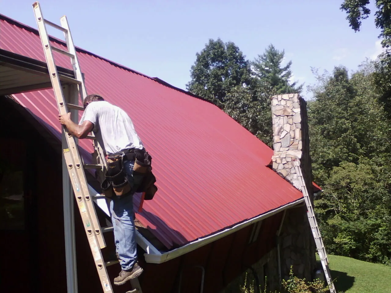 Roofer on a ladder beside a red metal roof and stone chimney — Soffit & Fascia Repair by Roofing Brothers in Coplay